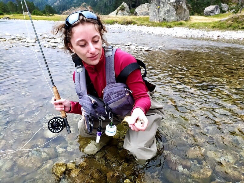 Femme en waders examinant un petit objet dans sa main, debout dans une rivière peu profonde et claire.