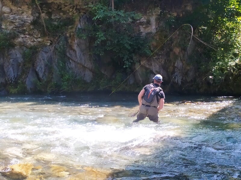 Pêcheur debout dans une rivière avec des rapides, lançant sa ligne sous un ciel partiellement nuageux.