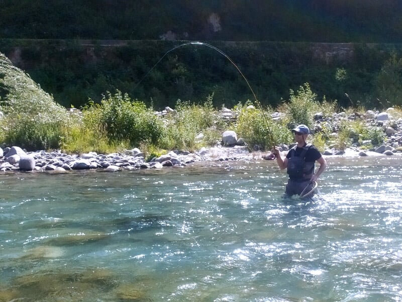 Pêcheur en waders dans une rivière large et peu profonde, lançant sa ligne avec des montagnes en arrière-plan.