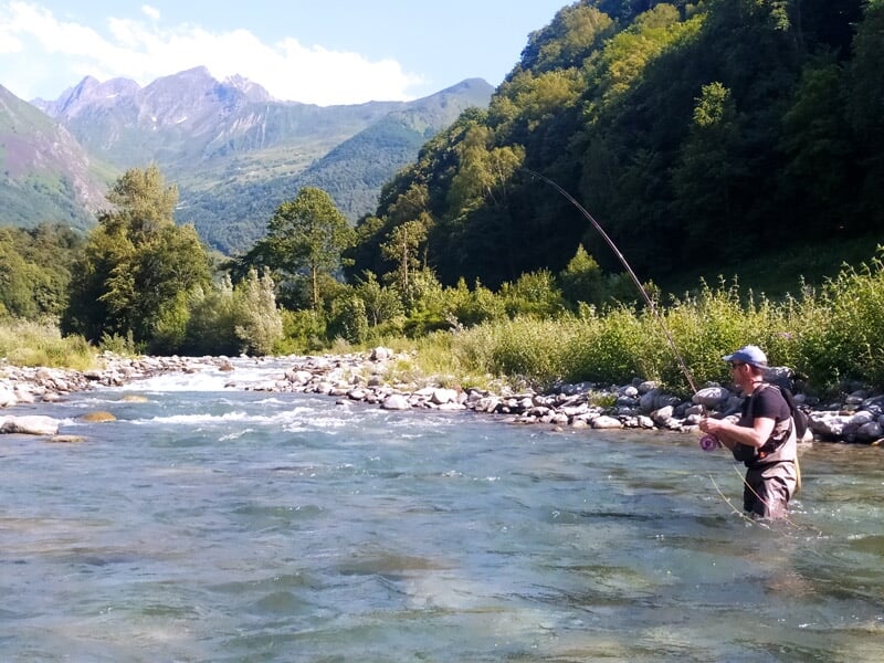 Pêcheur dans une rivière entourée de montagnes et de végétation, concentré sur sa ligne de pêche.