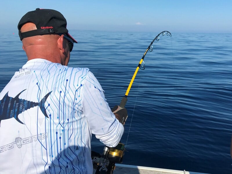 Homme en train de pêcher en mer avec une canne à pêche, portant une casquette et un t-shirt blanc à motif poisson.