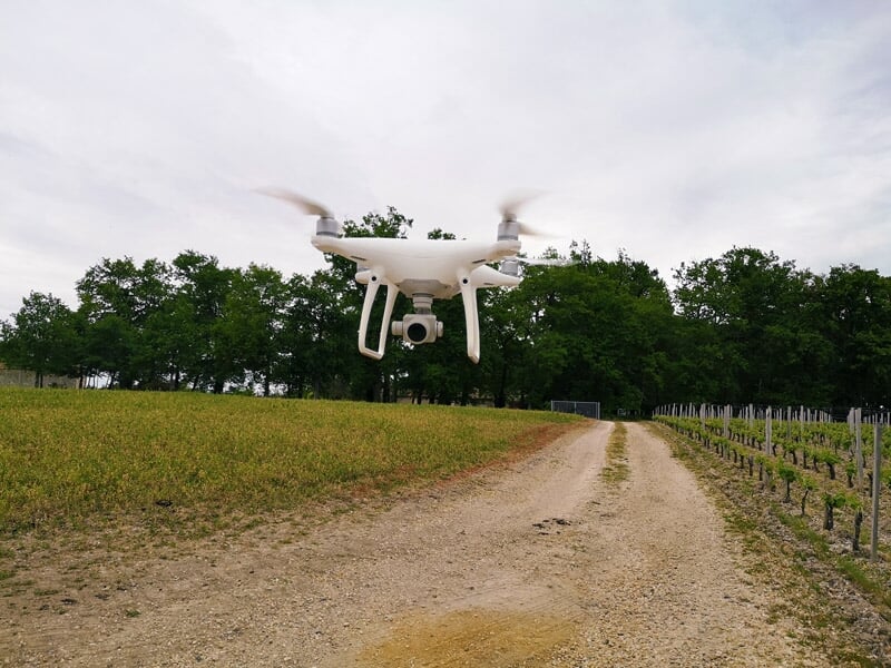 Drone blanc en vol stationnaire au-dessus d'un chemin de terre entouré de vignes et d'arbres.