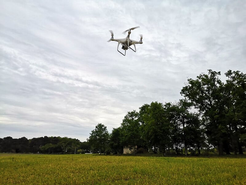 Drone blanc volant au-dessus d'un champ herbeux avec des arbres et un ciel nuageux en arrière-plan.