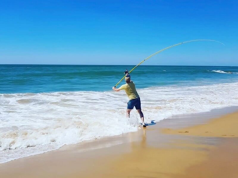 Homme lançant une ligne de pêche sur une plage ensoleillée avec l'océan et le ciel bleu en arrière-plan.
