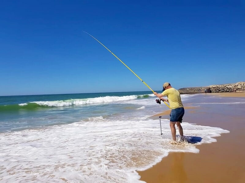 Pêcheur debout dans l'eau sur une plage, tenant une canne à pêche face à la mer sous un ciel clair.