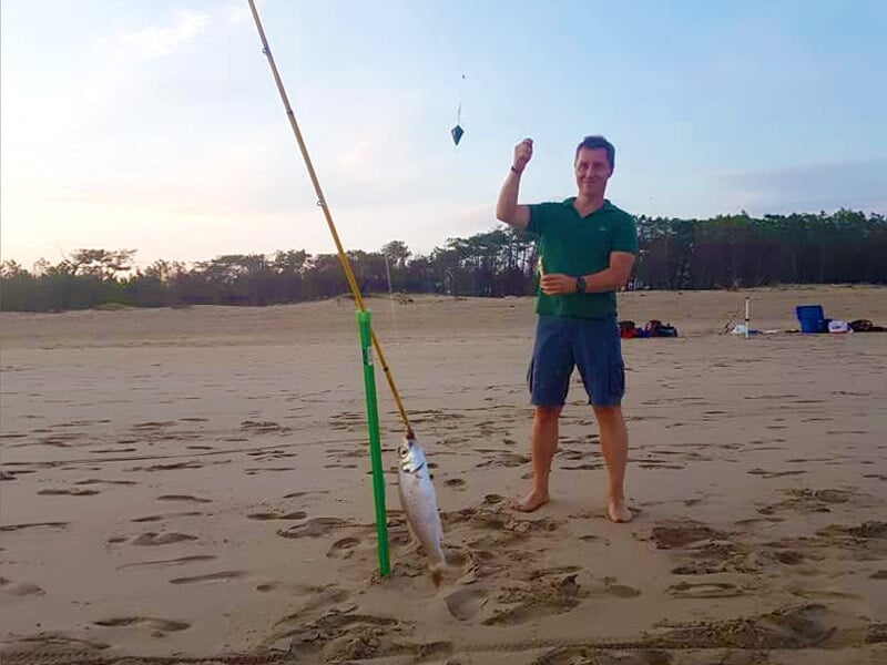 Homme souriant sur la plage tenant une canne à pêche avec un poisson accroché, sable et forêt en arrière-plan.