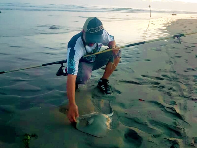 Pêcheur accroupi sur le sable tenant une raie capturée, canne à pêche posée dans le sable.