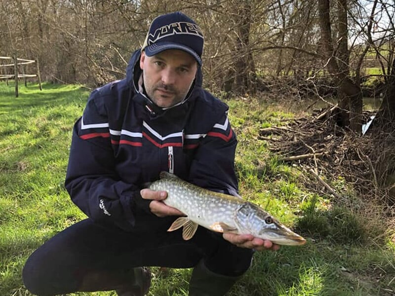 Homme en tenue de pêche tenant un brochet devant un décor de forêt et d'herbe verte.