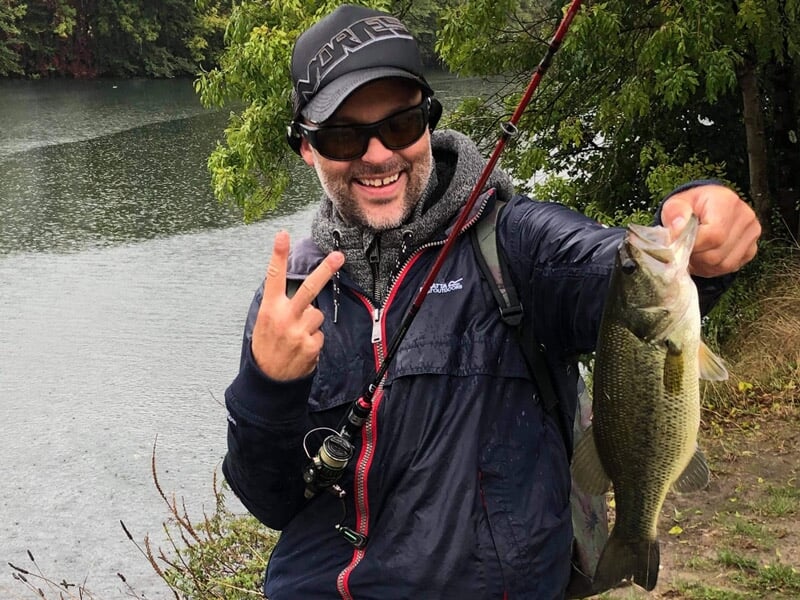 Pêcheur souriant tenant un gros poisson noir et blanc près d'un plan d'eau entouré d'arbres.