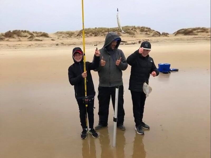 Trois personnes sur une plage, dont un enfant avec une canne à pêche, posant sous un ciel nuageux.