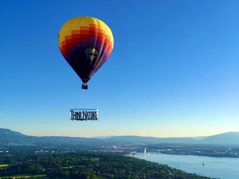 Montgolfière multicolore avec une bannière 'Think Nature' survolant un paysage urbain et lacustre.