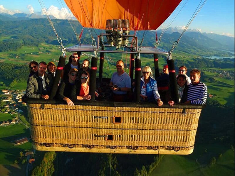 Groupe de personnes dans une nacelle de montgolfière survolant une vallée verdoyante et montagneuse.