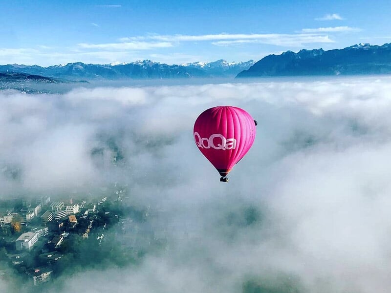 Montgolfière rose survolant une mer de nuages au-dessus d'une ville et des montagnes.
