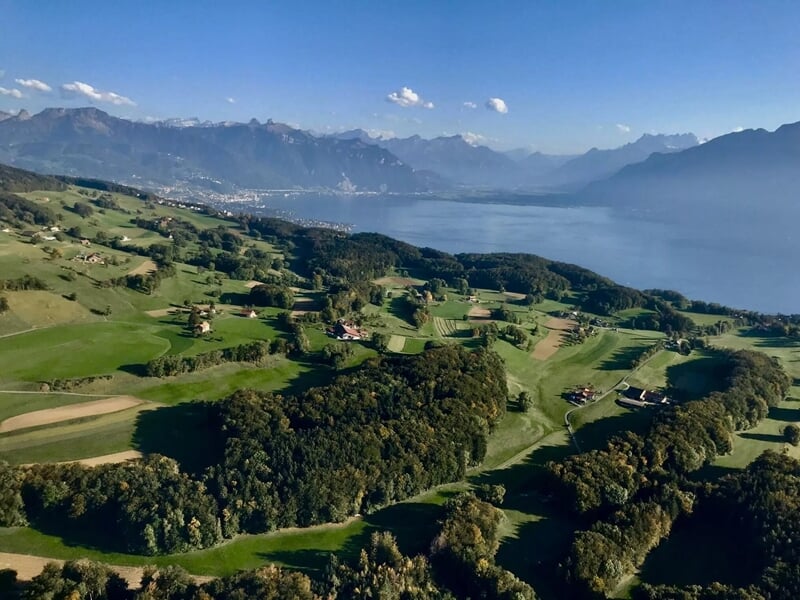 Paysage vallonné avec des champs verts et un grand lac sous un ciel bleu clair.