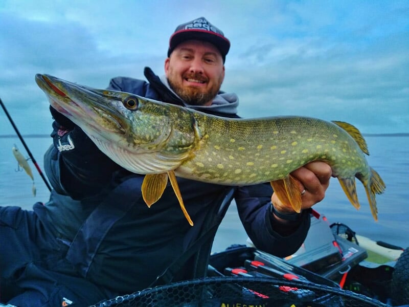 Homme souriant en tenue de pêche tenant un brochet dans un kayak sur un lac calme au lever du jour.