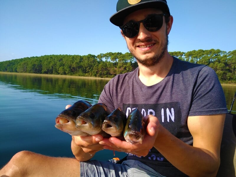 Jeune homme en t-shirt et lunettes de soleil montrant trois petits poissons pêchés, assis sur un kayak.