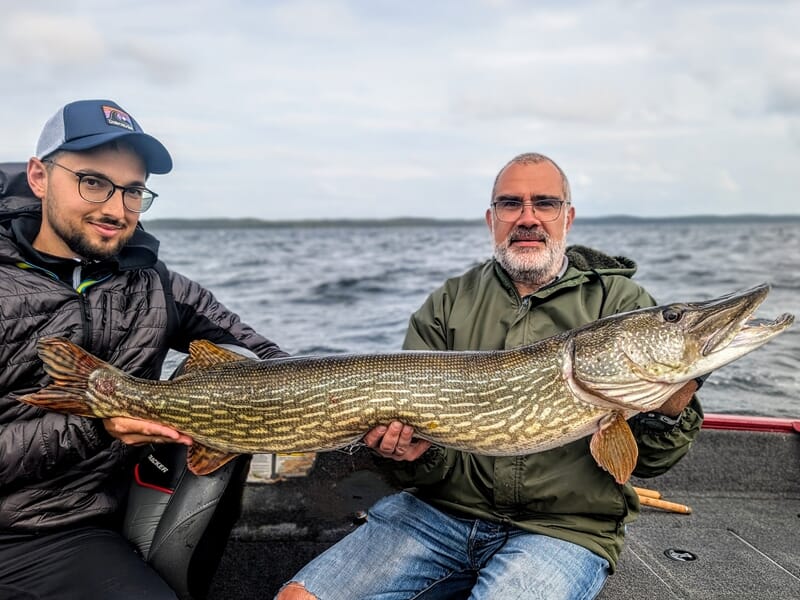 Deux hommes sur un bateau tenant un grand brochet fraîchement pêché sur un lac sous un ciel nuageux.