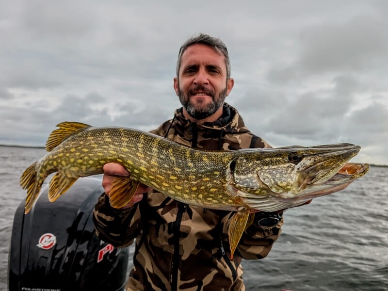 Un homme souriant en tenue camouflage tenant un brochet impressionnant sur un bateau en pleine eau.