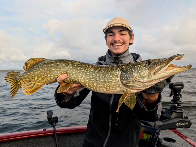 Un jeune homme souriant tenant un brochet de grande taille sur un bateau au milieu d'un lac.