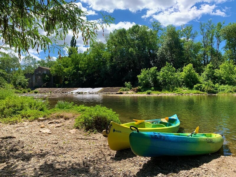 Deux kayaks colorés posés sur la rive d'une rivière calme entourée d'arbres verts sous un ciel bleu.