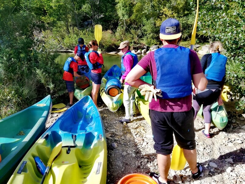 Groupe de personnes en tenue de sport et gilets de sauvetage préparant des kayaks au bord d'une rivière.