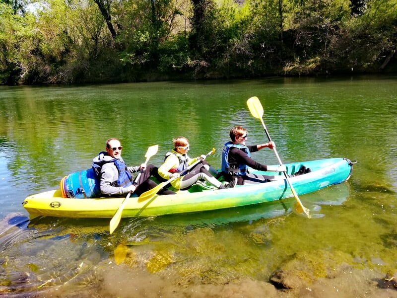 Trois personnes pagayant dans un kayak sur une rivière claire avec des rochers visibles sous l'eau.
