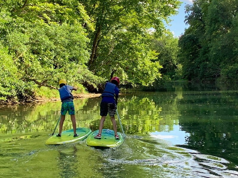 Deux enfants debout sur des planches de stand-up paddle pagayant sur une rivière calme entourée d'arbres.