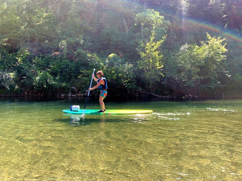 Personne debout sur une planche de stand-up paddle pagayant sur une rivière claire sous un ciel ensoleillé.