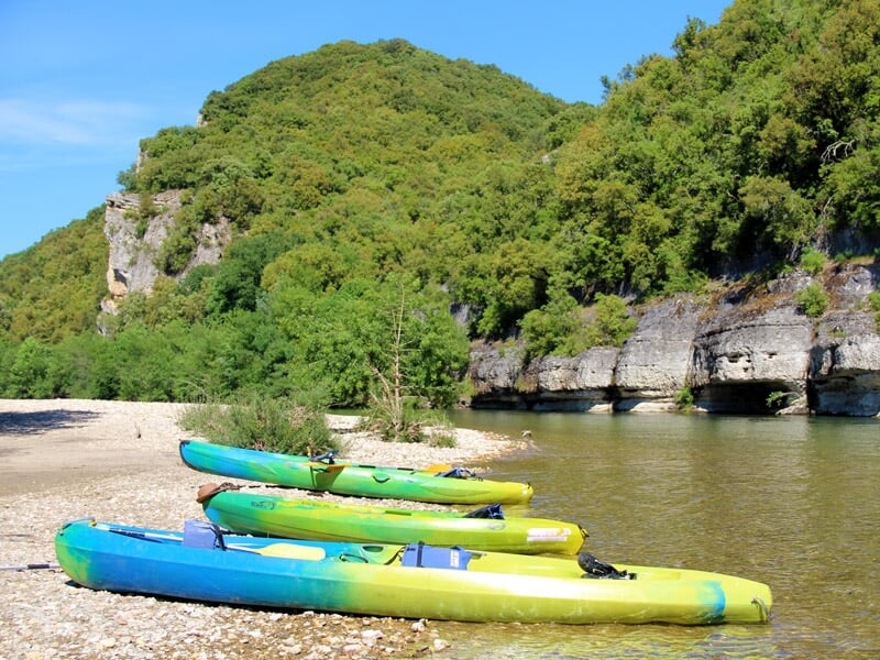 Deux personnes pagayant dans un kayak bleu et vert sur une rivière calme bordée d'arbres et de rochers.
