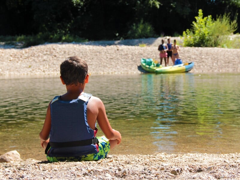 Plusieurs kayaks colorés alignés sur une plage de galets au bord d'une rivière entourée de collines boisées.