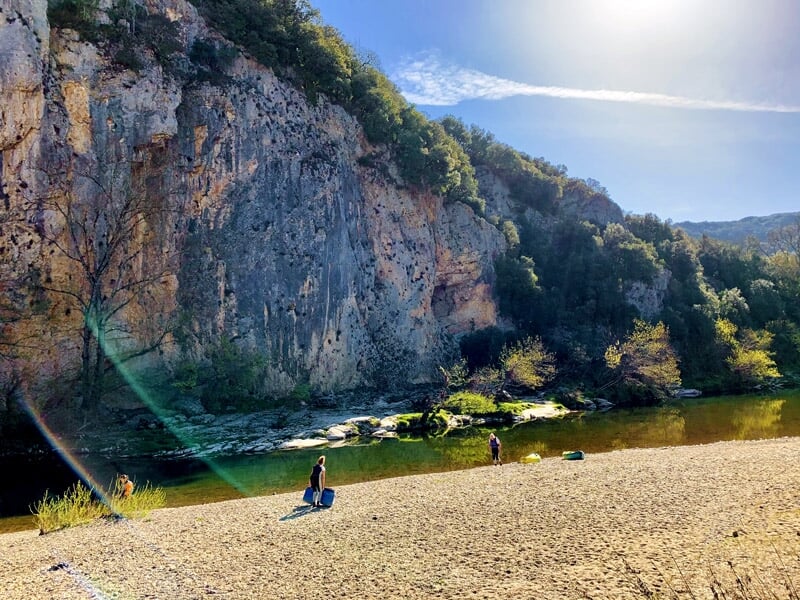 Enfant assis sur une plage de galets regardant deux personnes pagayer sur un kayak au bord d'une rivière.