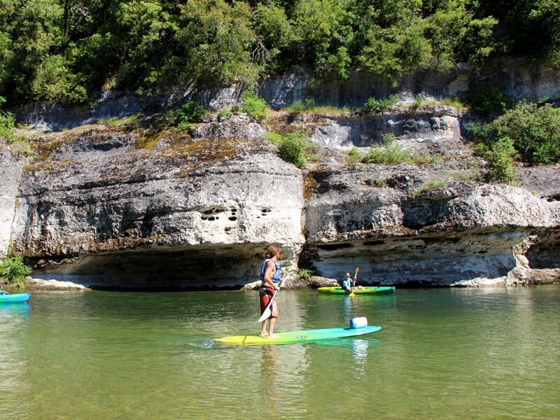 Deux enfants debout sur des planches de stand-up paddle pagayant sur une rivière calme entourée d'arbres verts.