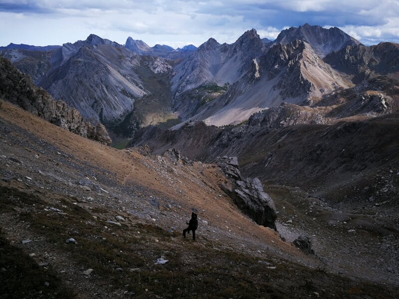 Un chien noir seul sur un sentier de montagne rocheuse avec des sommets escarpés en arrière-plan sous un ciel nuageux.