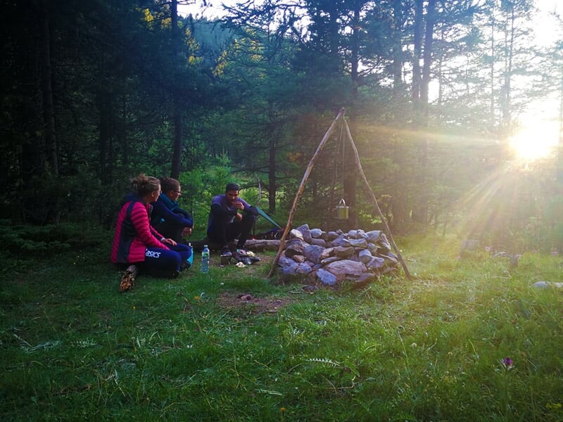 Groupe de personnes assises sur l'herbe près d'un feu de camp entouré de pierres dans une clairière en forêt au soleil couchant.