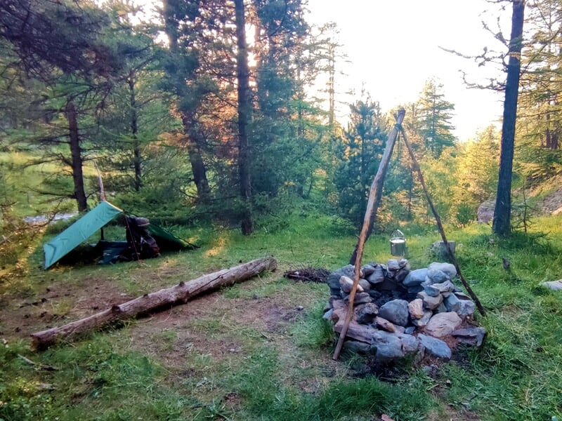 Campement en forêt avec un feu de camp entouré de pierres, un abri de fortune en bâche verte et des troncs d'arbres posés au sol.