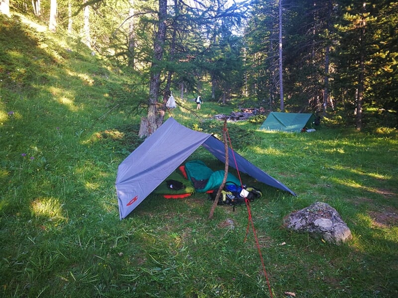 Tarp gris tendu en forêt servant d'abri, avec des sacs et du matériel de camping posés sur l'herbe autour.