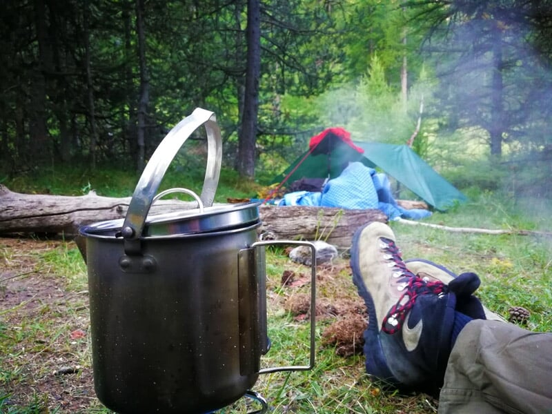 Gros plan d'une casserole métallique posée près d'un feu de camp, avec une personne assise en arrière-plan portant des chaussures de randonnée.