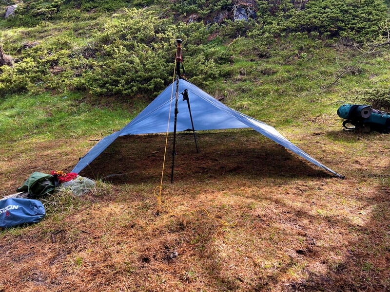 Tarp bleu tendu en forêt sur un sol herbeux, avec des bâtons de randonnée et un sac à dos posés à côté.
