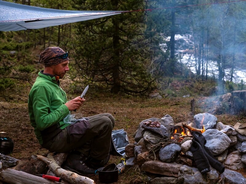 Homme assis près d'un feu de camp en forêt, tenant un couteau et préparant quelque chose, avec de la fumée autour.