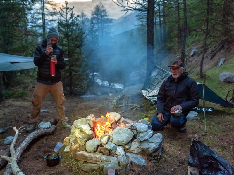 Deux hommes assis près d'un feu de camp en forêt, entourés de pierres, avec des arbres et des bâches en arrière-plan.