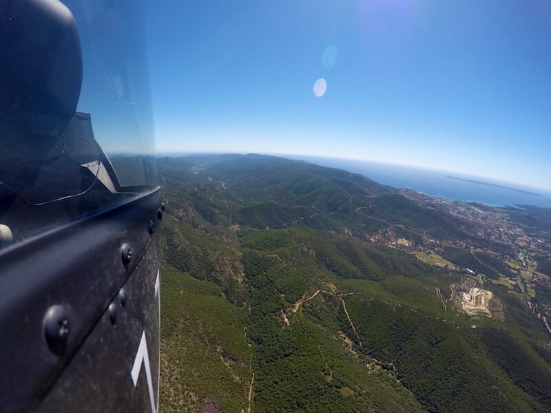 Vue aérienne d'une chaîne de montagnes verdoyantes s'étendant jusqu'à la mer Méditerranée. Les collines et les vallées sont couvertes de forêts denses, avec des sentiers serpentant à travers le paysag