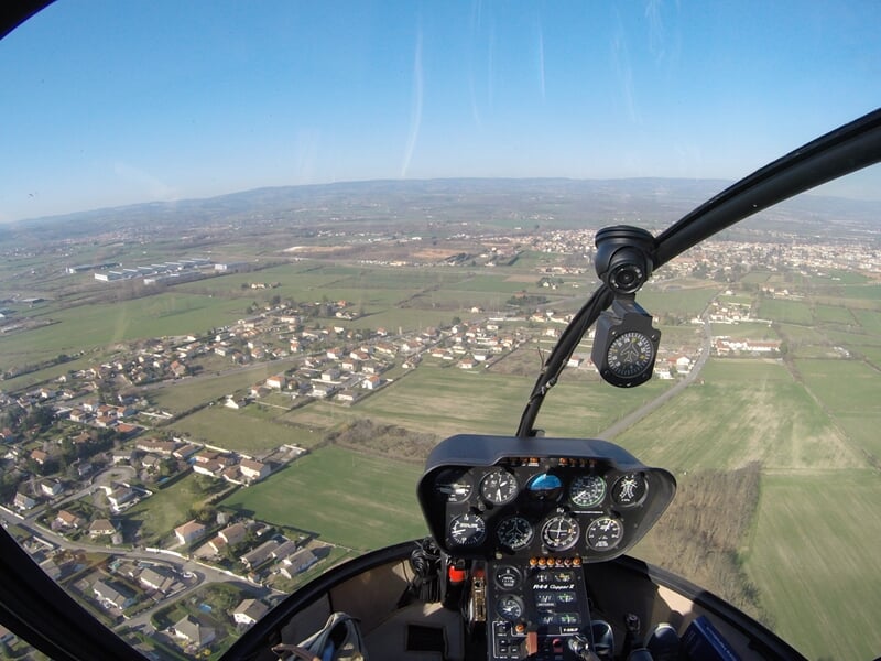Cockpit d'un hélicoptère en vol au-dessus d'une campagne avec champs et maisons dispersées, lors d'un baptême dans le Forez.