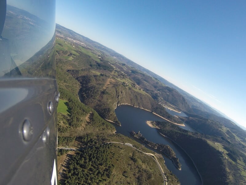 Paysage vu d'un hélicoptère montrant un lac entouré de collines verdoyantes et routes sinueuses.