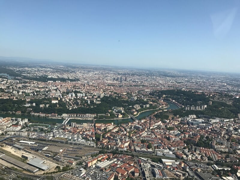 Vue aérienne de Lyon avec ses bâtiments, ses quartiers denses et les collines environnantes. La Saône serpente à travers la ville, traversant des zones urbaines et verdoyantes