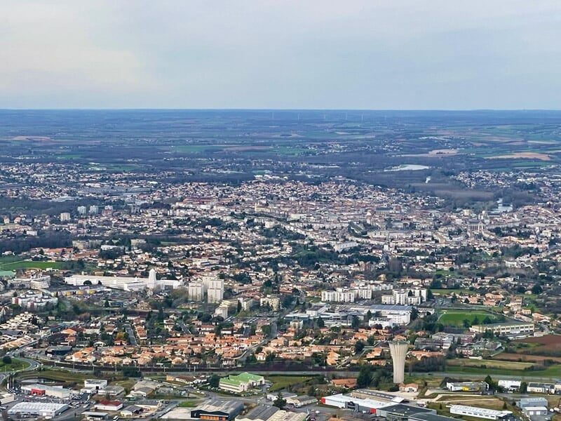 Vue aérienne d'une ville avec toits rouges et rues sous un ciel clair et lumineux.