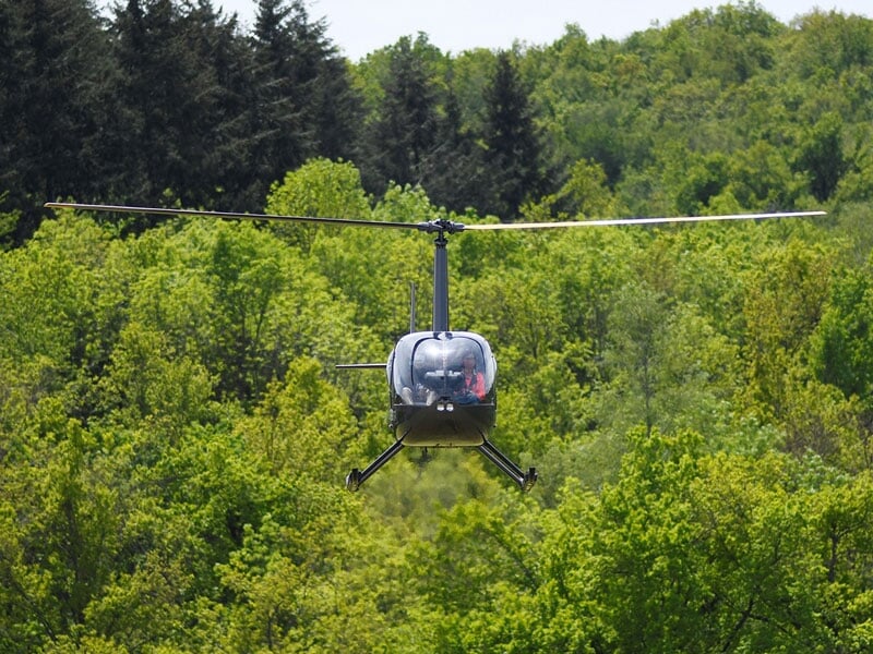 Hélicoptère noir en vol stationnaire au-dessus d'une forêt dense et verdoyante par une journée ensoleillée.
