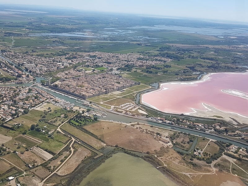 Vue aérienne de la ville fortifiée de Aigues-Mortes avec ses remparts, un canal et un lac salé rose à côté.