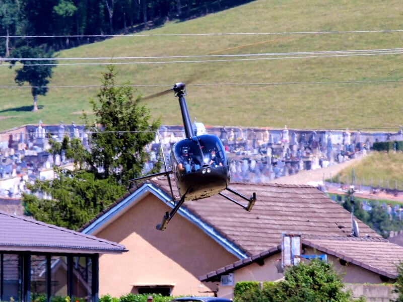 Un hélicoptère noir en vol bas au-dessus de toits de maisons avec un paysage de collines et arbres en arrière-plan.