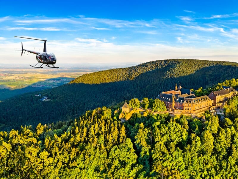Hélicoptère en vol près d'une abbaye historique nichée dans une forêt dense sur une colline, avec un panorama sur des vallées lointaines sous un ciel bleu en fin de journée