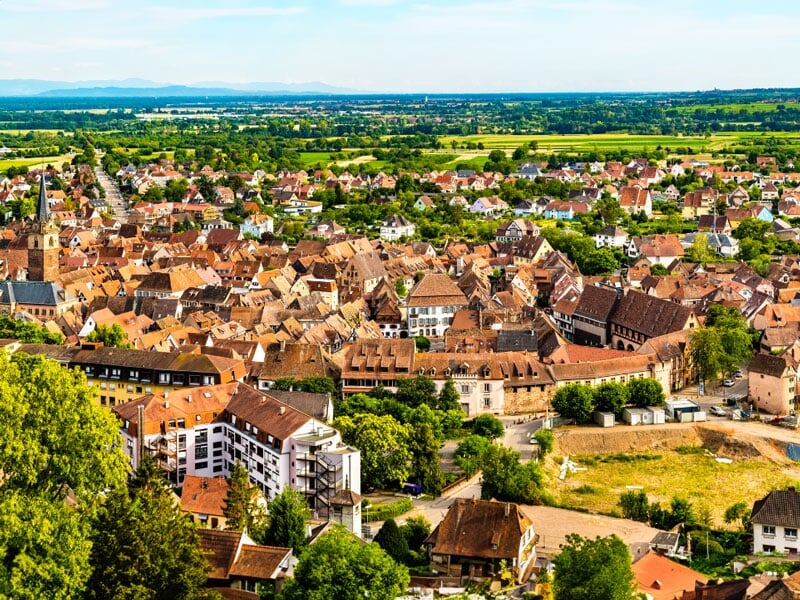 Paysage urbain dense d'une ville historique avec des toits en tuiles rouges, des clochers d'églises et des rues organisées en quadrillage, entourée de champs agricoles et d'une périphérie moderne